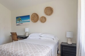 Une chambre d’hôtel comprenant un lit, deux paniers fixés au mur, un bureau avec une chaise, une lampe et un tableau représentant une scène de plage.