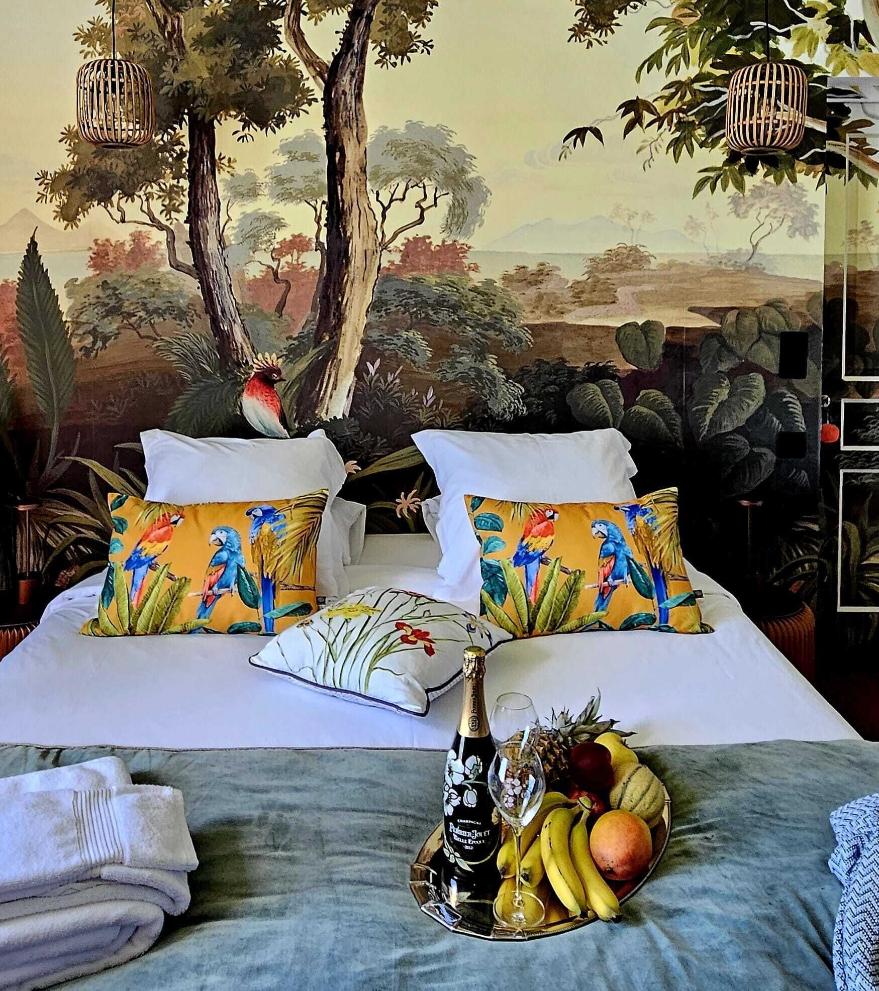 Una cama con dos almohadas mostrando aves tropicales, una botella de champán y una cesta de frutas sobre la mesita de noche.