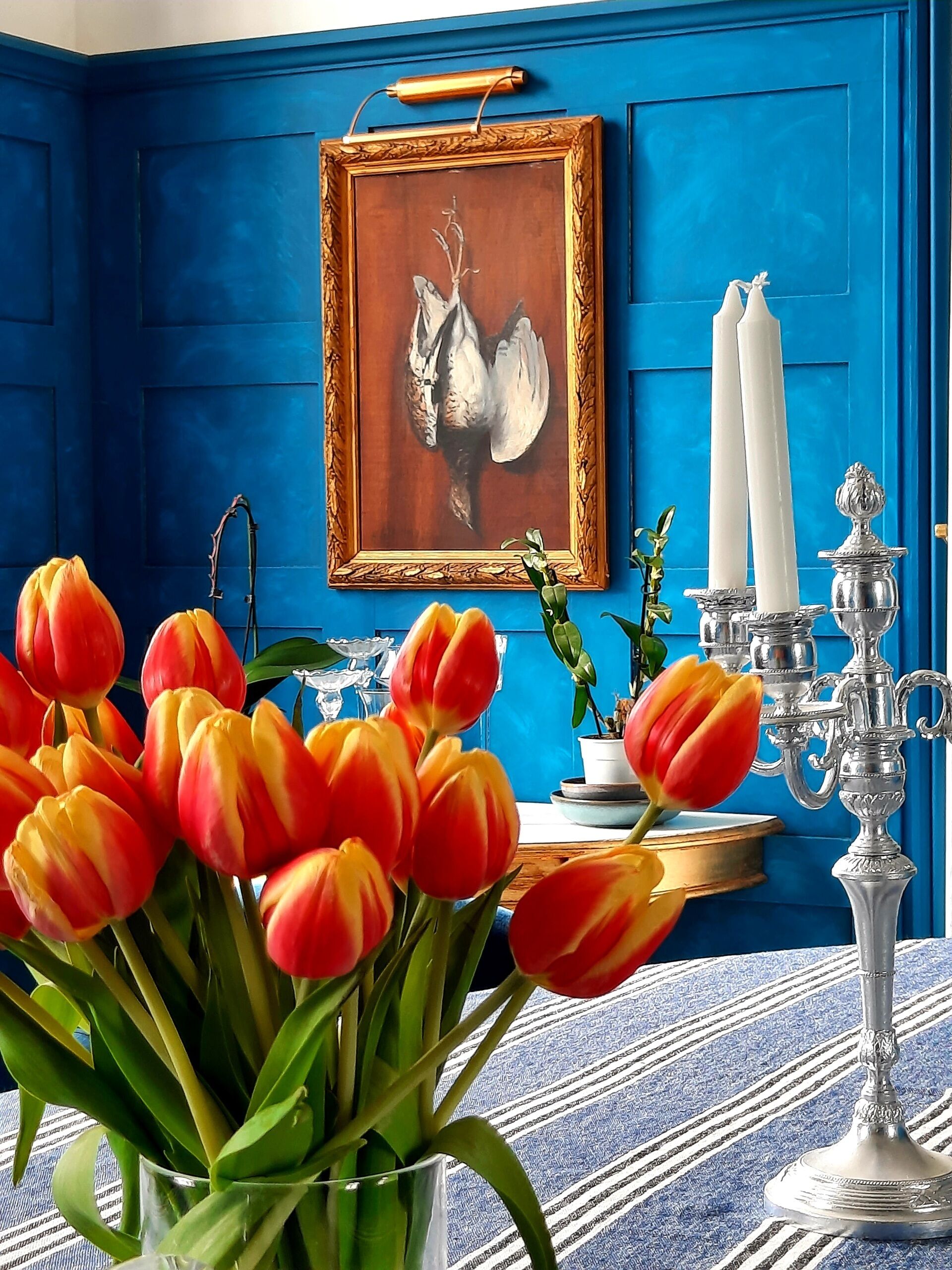 A room with blue walls, a framed painting, a vase of tulips, a candelabra, and a small table with a plant.