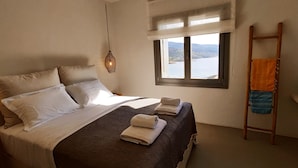 Un dormitorio con cama, dos almohadas, una ventana con vistas, una lámpara colgante y una escalera de madera.