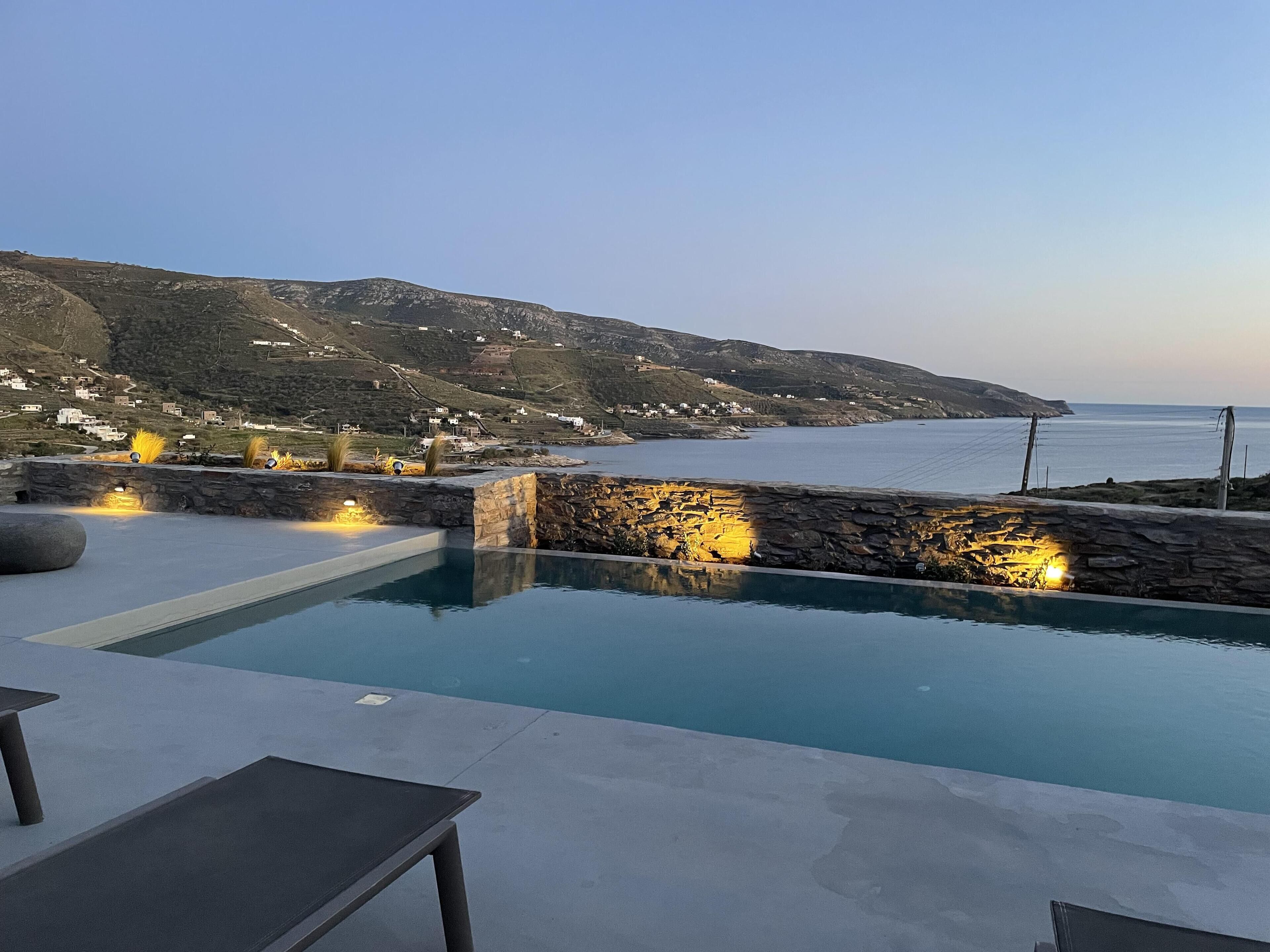 A pool area with stone walls and outdoor lighting, overlooking a coastal landscape with houses and hills.