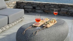 Una mesa de piedra con una silla de mimbre, dos copas de vino tinto y una tabla de madera con queso y embutidos. En el fondo, una pared de piedra y vistas al mar y colinas.