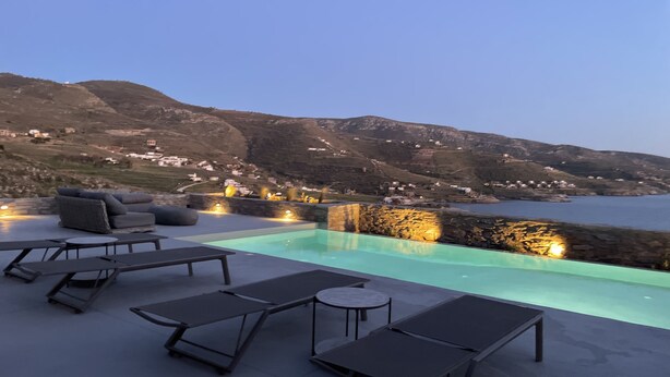 Una piscina infinita con sillas y mesas de descanso, con vistas a un paisaje costero con colinas y una masa de agua.