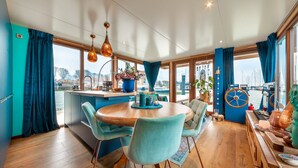 Un espace de repas moderne avec une table ronde en bois, des chaises de couleur turquoise et une vue sur l’eau grâce à de grandes fenêtres.