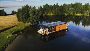 Une péniche dotée d’un toit recouvert de panneaux solaires est amarrée sur un lac paisible, entourée d’une forêt dense.
