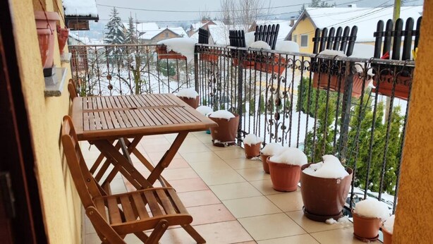 A balcony with a wooden table and chairs, potted plants, and a view of snow-covered houses. - Vila Domm in Rakovica (Rakovica)