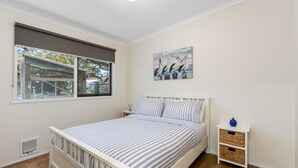 A bedroom with a bed, a window, a wall-mounted painting, a nightstand, and a small table.