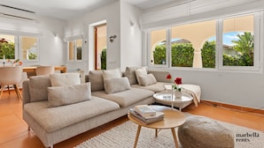 A modern living room with a beige sectional sofa, a round coffee table, and a view of a garden through large windows. - Beachfront Villa Casa Mosa | Ref. #32 (Marbella)
