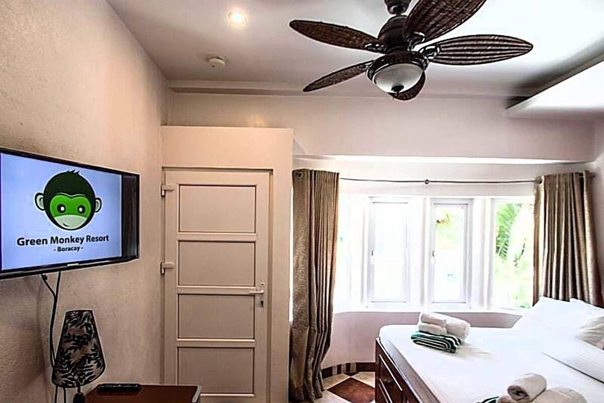 Habitación de hotel con cama, televisión mostrando 'Green Monkey Resort - Boracay', ventilador de techo y ventana con cortinas.