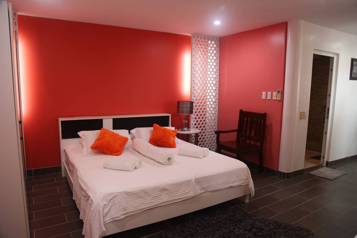 Habitación de hotel con una cama grande, ropa de cama blanca y almohadas naranjas. Hay una mesita con una lámpara y una silla. Las paredes son rojas y una puerta conduce a otra habitación.