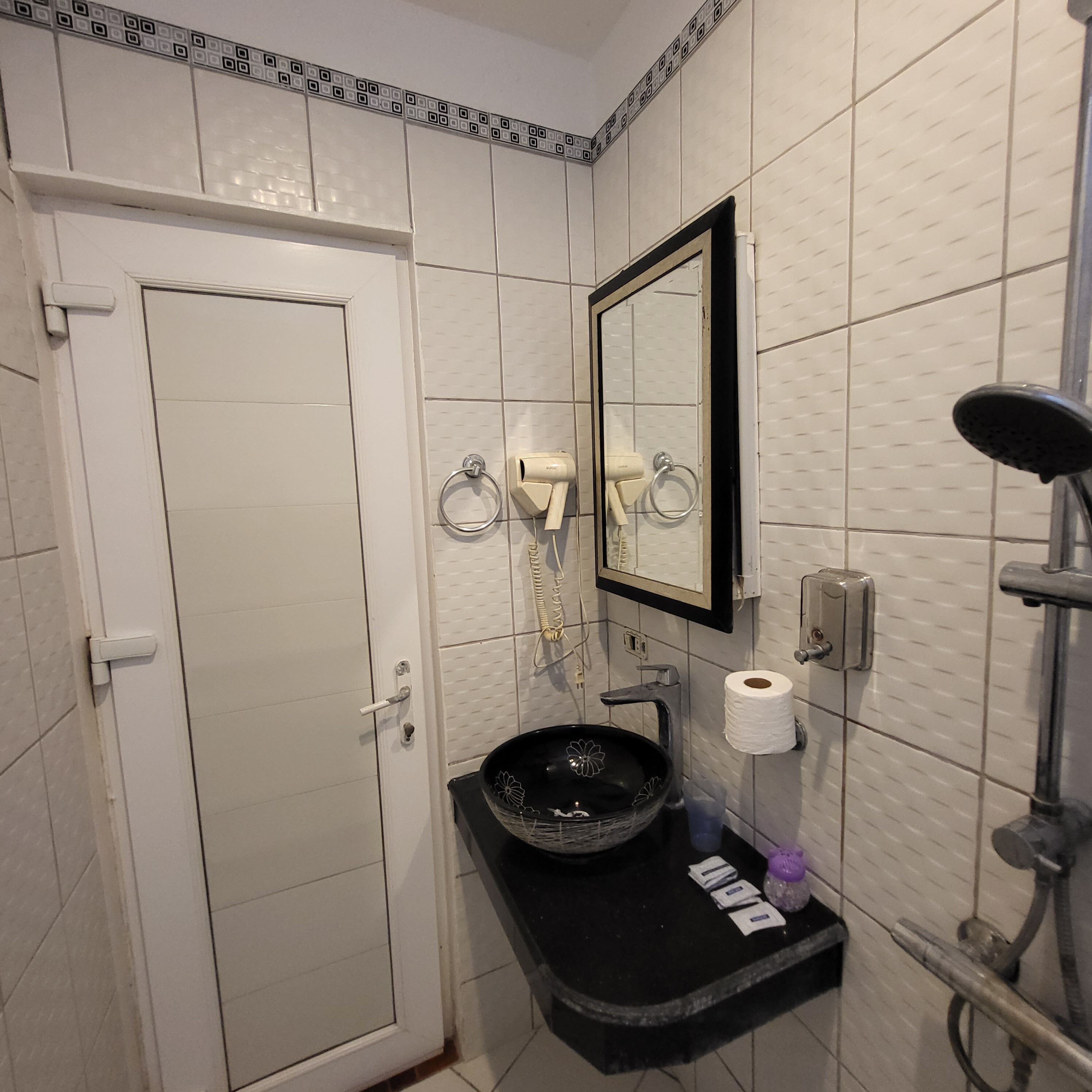 A bathroom with white tiled walls, a black sink, a mirror, a toilet paper holder, and a shower.