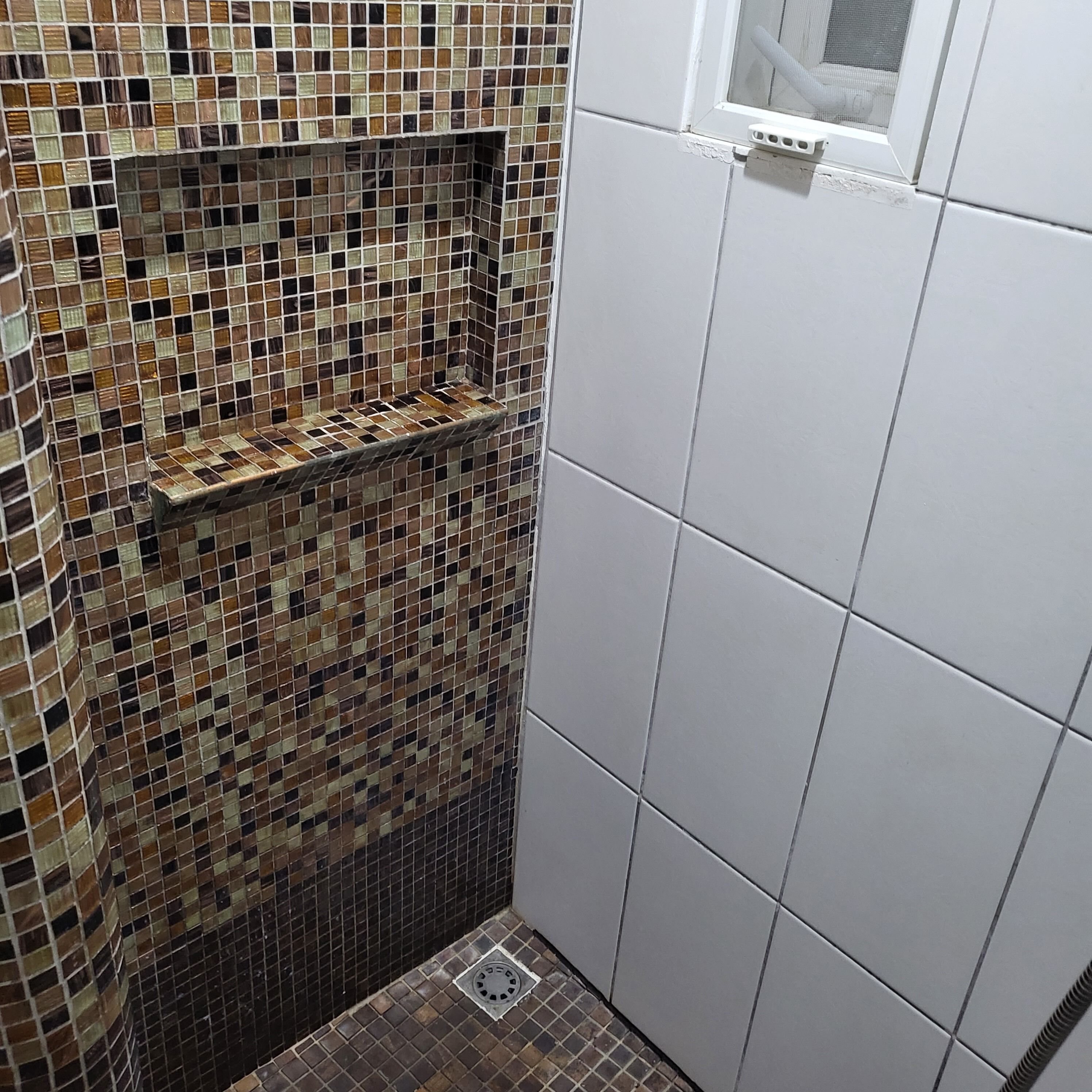 A bathroom with a mosaic tile wall, a white tiled shower area, and a window with a mesh screen.