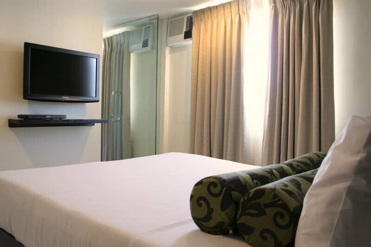 A hotel room with a bed, a TV mounted on the wall, a small shelf, and a window with curtains.