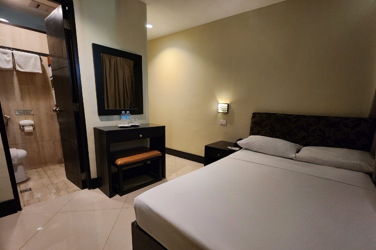 A hotel room with a bed, a desk with a chair, a nightstand, a mirror, a wall-mounted light, and a toilet.