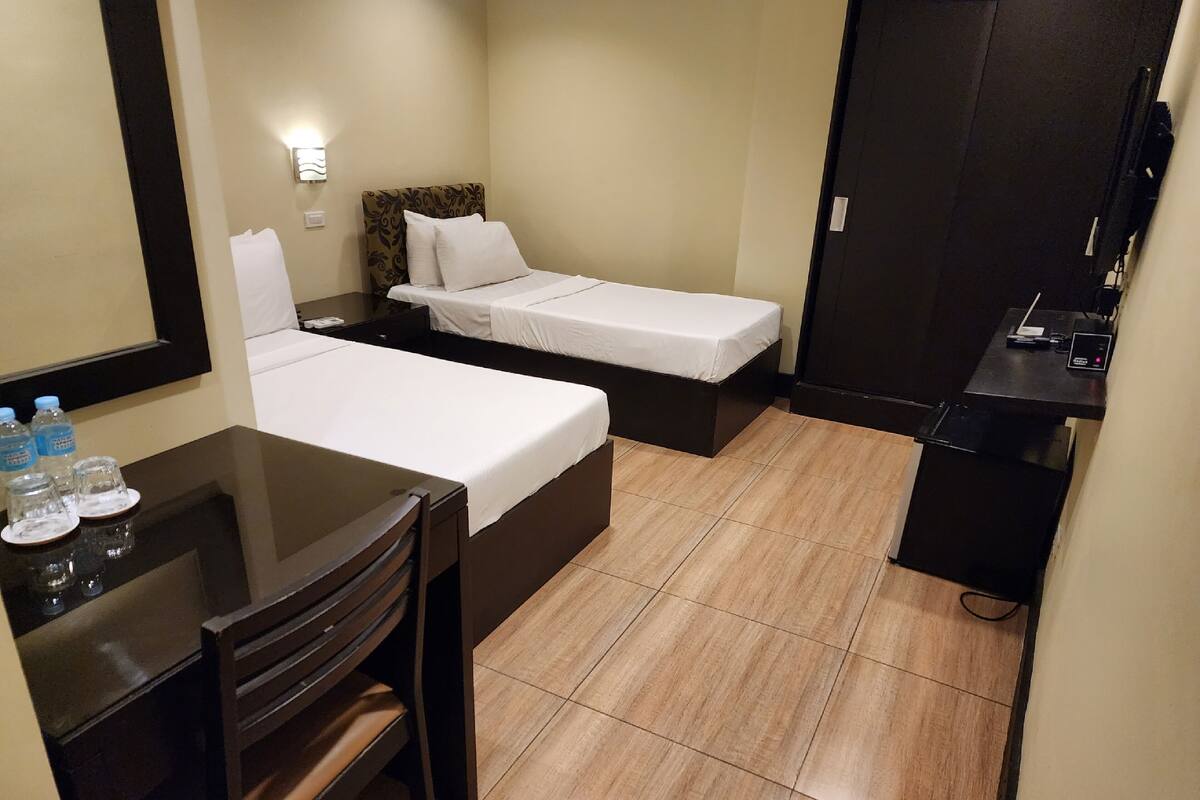 A hotel room with two single beds, a desk with a chair, a mirror, and a small table with water bottles.