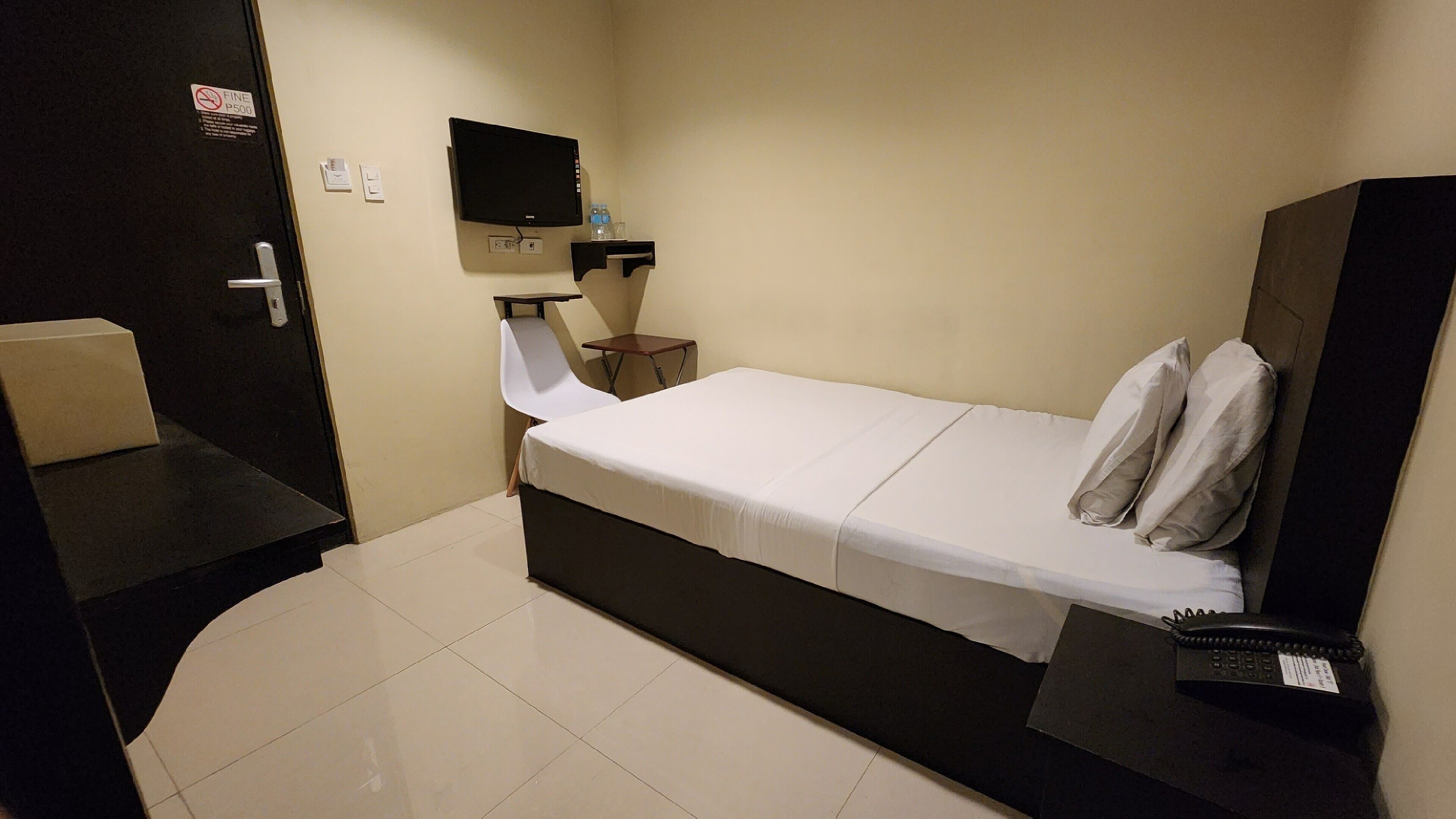 Een kleine, functionele hotelkamer met een bed, een bureau, een stoel, een aan de muur bevestigde televisie en een telefoon op het bureau.