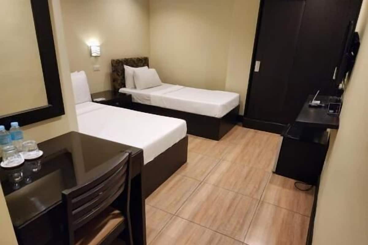 A small hotel room with two beds, a desk, and a chair. There is a mirror on the wall and a door to the right.