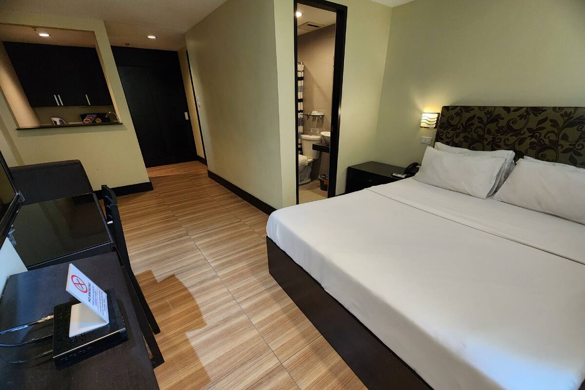 A hotel room with a large bed, a desk with a computer, a mirror, and a bathroom visible through an open door.