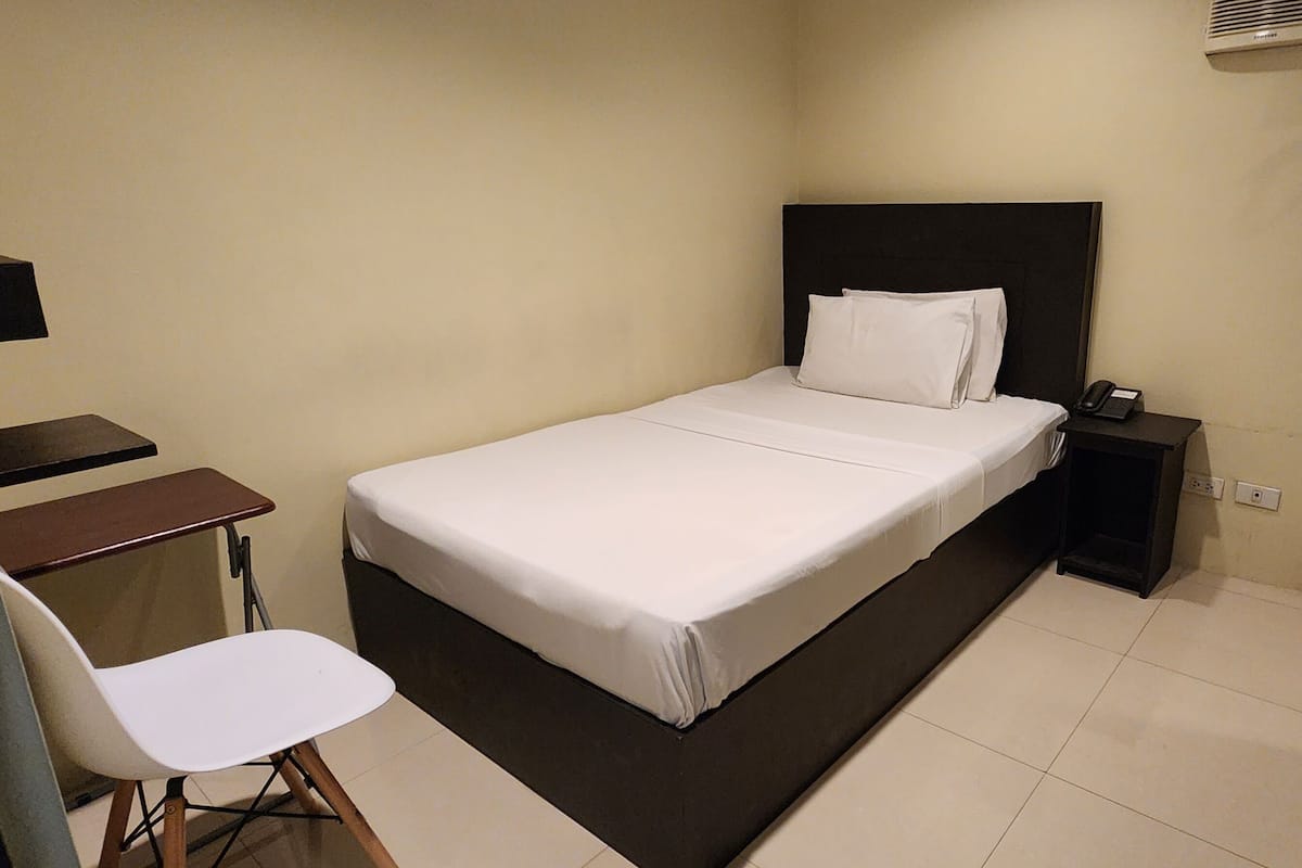 A bedroom with a bed, a desk, a chair, and a nightstand with a phone.