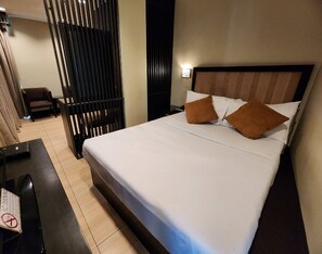 Una cama bien tendida con sábanas blancas y dos almohadas marrones, una mesita de noche negra con una lámpara, y una silla en una habitación de hotel.
