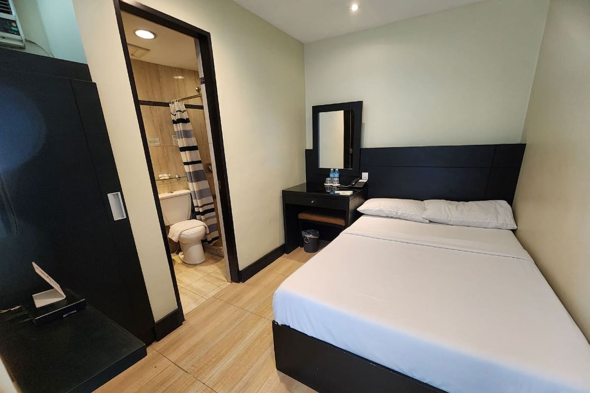 A hotel room with a bed, a bedside table, a mirror, a small desk, a toilet, and a shower area.