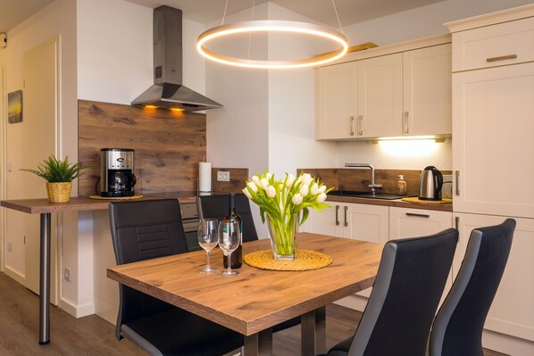 A modern kitchen with a wooden dining table, black chairs, a coffee machine, and a vase with white flowers.