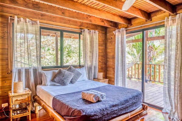A wooden cabin bedroom with a large bed, two bedside tables, and a view of the outdoors through a sliding glass door. - Rustic mountain-view home near the beach with private pool, deck, W/D, & WiFi (Dominical)