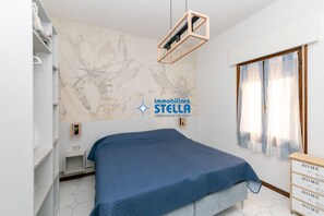 A bedroom with a bed, a wall-mounted light, a window with curtains, and a wall with a floral design.