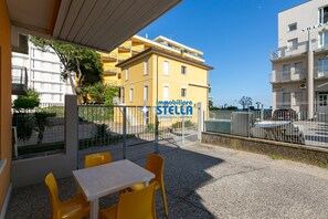 An outdoor seating area with yellow chairs and a table, a building with balconies, and a clear blue sky. - * Giglio * / Piazza Marconi [park] (Lido di Jesolo)