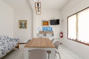 A room with a wooden table, chairs, a bed with floral bedding, a mounted TV, and a fire extinguisher.