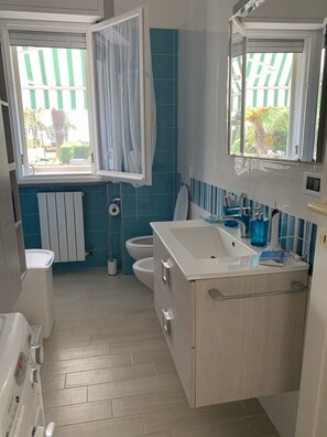 A modern bathroom with a white sink, a toilet, and a bathtub. There are windows with curtains, a radiator, and a mirror above the sink. - Relax right by the sea in Silvi Marina.<br>Jun 25–Jul 30–Aug 20 (Silvi Marina)