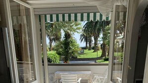 Una stanza luminosa con una porta a vetri scorrevoli che dà su un balcone con tavolo e sedie, un piccolo tavolino con una lampada e una presa di corrente a pavimento.