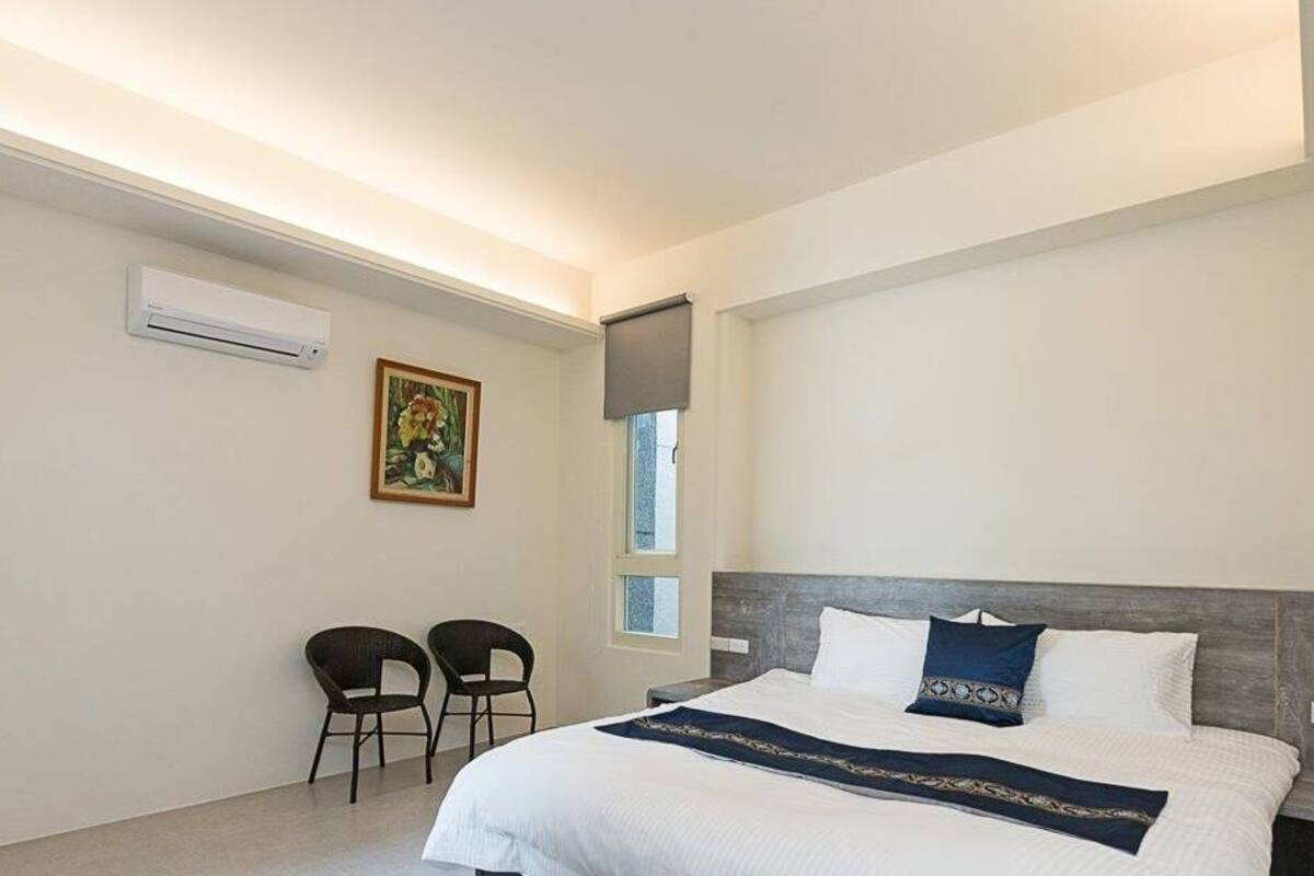 A modern hotel room with a large bed, two chairs, an air conditioning unit, a window, and a painting on the wall.