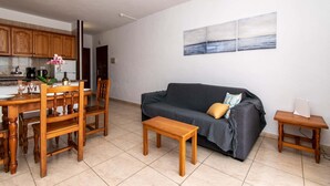 Un salon avec un canapé gris foncé, une table basse en bois et une petite table d’appoint en bois. On y trouve également un coin cuisine avec des placards en bois et une table à manger avec des chaises.