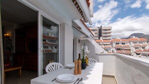 An outdoor dining area with a white table set for two, a glass door leading to a kitchen, and a view of a residential area with red-tiled roofs. - Las Blanquitas 9 (Playa de Las Americas)