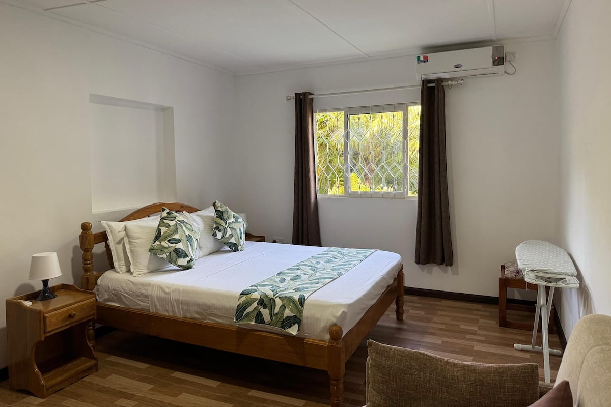 A bedroom with a wooden bed, a nightstand, a chair, a window with curtains, and an air conditioning unit.