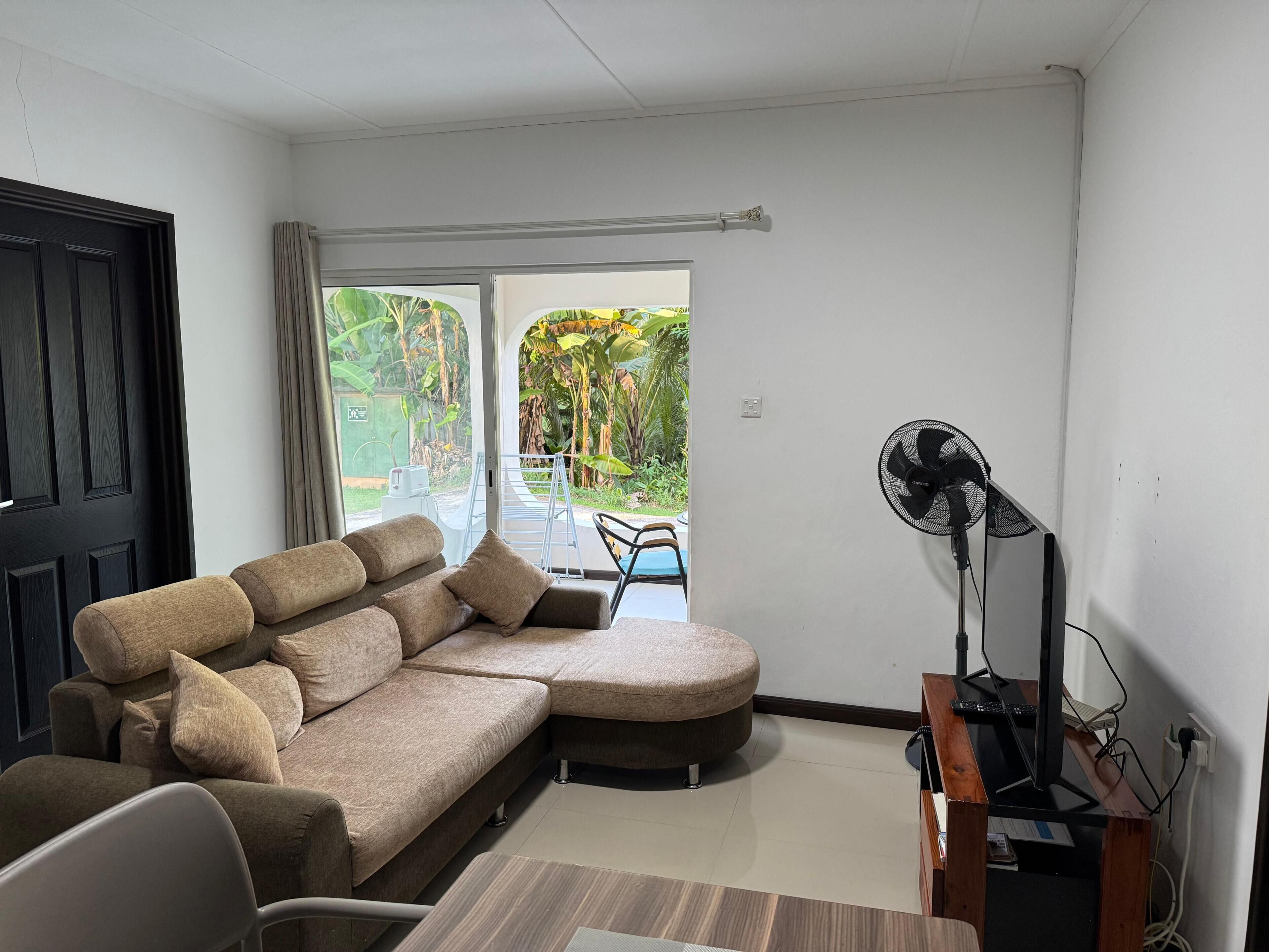 A modern living room with a sectional sofa, a desk with a computer, a standing fan, and a view of a garden through a sliding door.