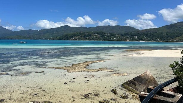 A beach with clear turquoise waters, a boat in the distance, and a mountainous backdrop. - Paul's Residence (Mahé Island)