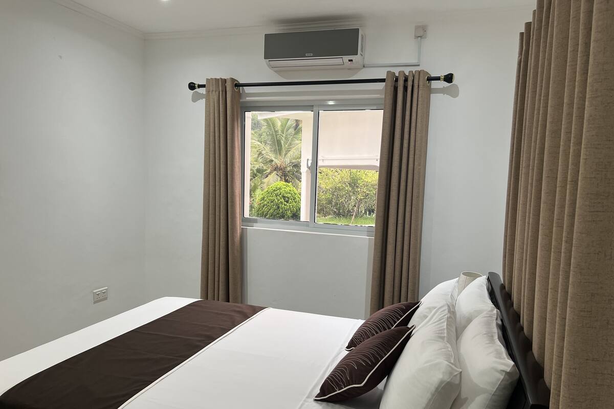 A hotel room with a bed, a window with curtains, an air conditioning unit, and a view of greenery outside.