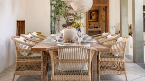 A dining area with a round table set for six, surrounded by wicker chairs, white paper lanterns hanging from the ceiling, and a vase with yellow flowers on the table.
