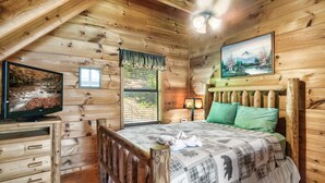 Une chambre confortable de style cabane, avec un intérieur en bois, un lit recouvert d’une couverture à carreaux, un téléviseur posé sur une commode en bois et un tableau accroché au mur.