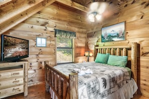 Um quarto de cabana acolhedor com decoração em madeira, uma cama com colcha xadrez, uma televisão sobre uma cômoda de madeira e um quadro na parede.