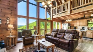 Un salon spacieux avec un grand canapé en cuir marron, une table basse en bois et un tapis à motifs. On y trouve deux fauteuils et une petite table avec des lampes. La pièce est dotée de grandes fenêtres qui offrent une vue sur une zone boisée.