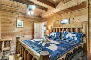 A wooden cabin interior with a bed featuring a blue and white quilt, a wooden headboard, and a ceiling fan.