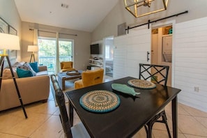 A modern living room with a sofa, coffee table, and dining table. There is a TV mounted on the wall, and a laundry room is visible in the background. - 2 Adult Bikes! Walk to Pool & Beach! Heart of Seagrove - Summer Breeze at Seagrove Beach 30A (Santa Rosa Beach)