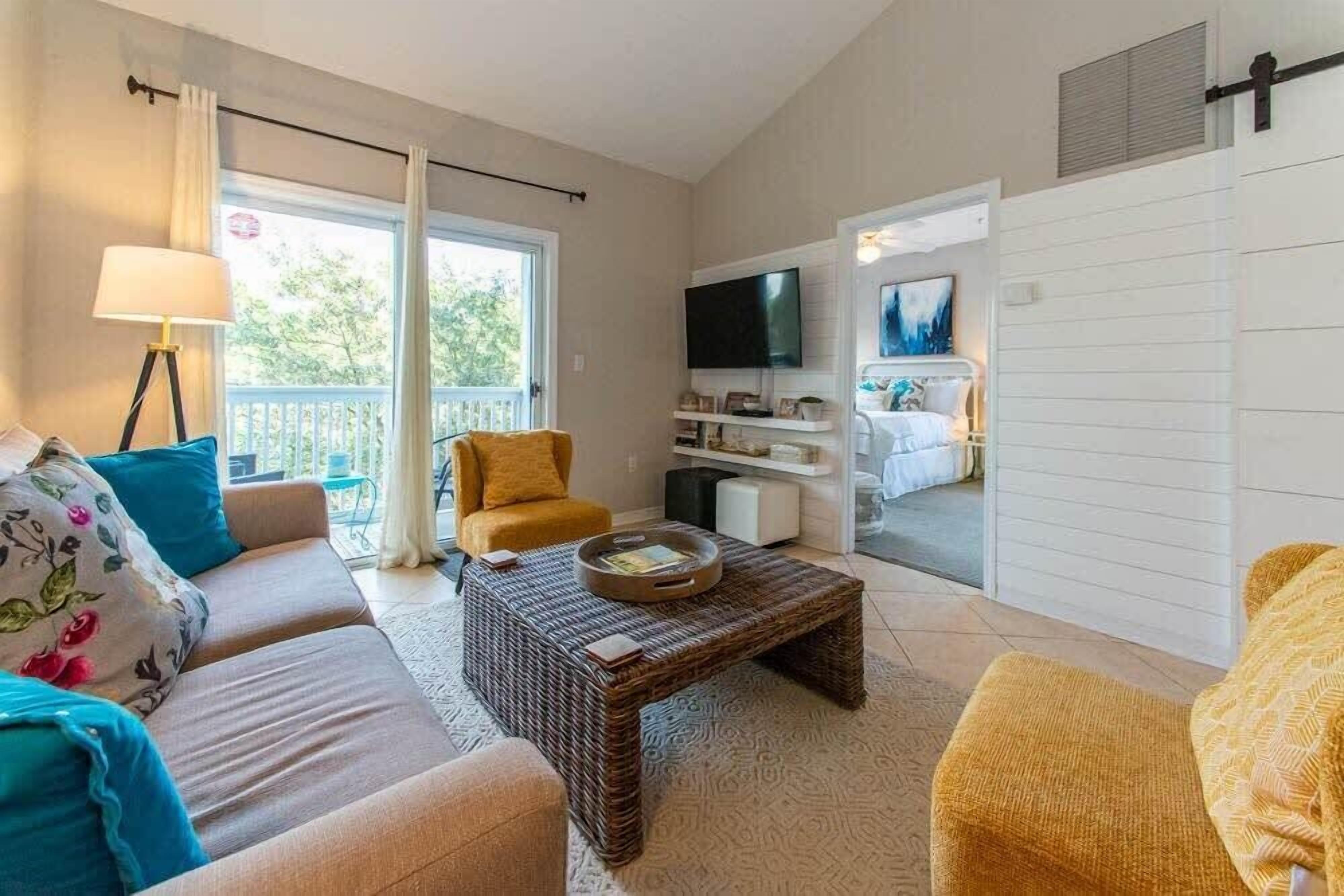 Een woonkamer met een bank, fauteuil en salontafel. Er is een televisie aan de muur bevestigd en een raam met gordijnen.