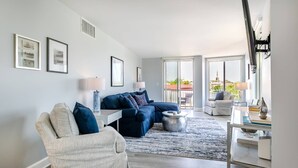 A modern living room with a sectional sofa, a white armchair, and a coffee table. There are framed pictures on the wall, a lamp, and a view of the city through large windows. - Prime Eastport Location - Waterfront Condo w Pool & Parking (Annapolis)