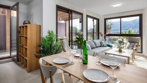 Ein Essbereich mit einem Holztisch, gedeckt für vier Personen, umgeben von Stühlen. Ein Sofa und ein kleiner Tisch mit einer Pflanze sind ebenfalls vorhanden. Große Fenster bieten Blick ins Freie.