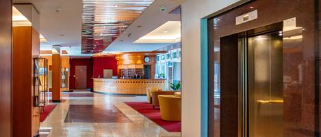 A hotel lobby with a marble floor, a reception desk, a red carpet, and an elevator.