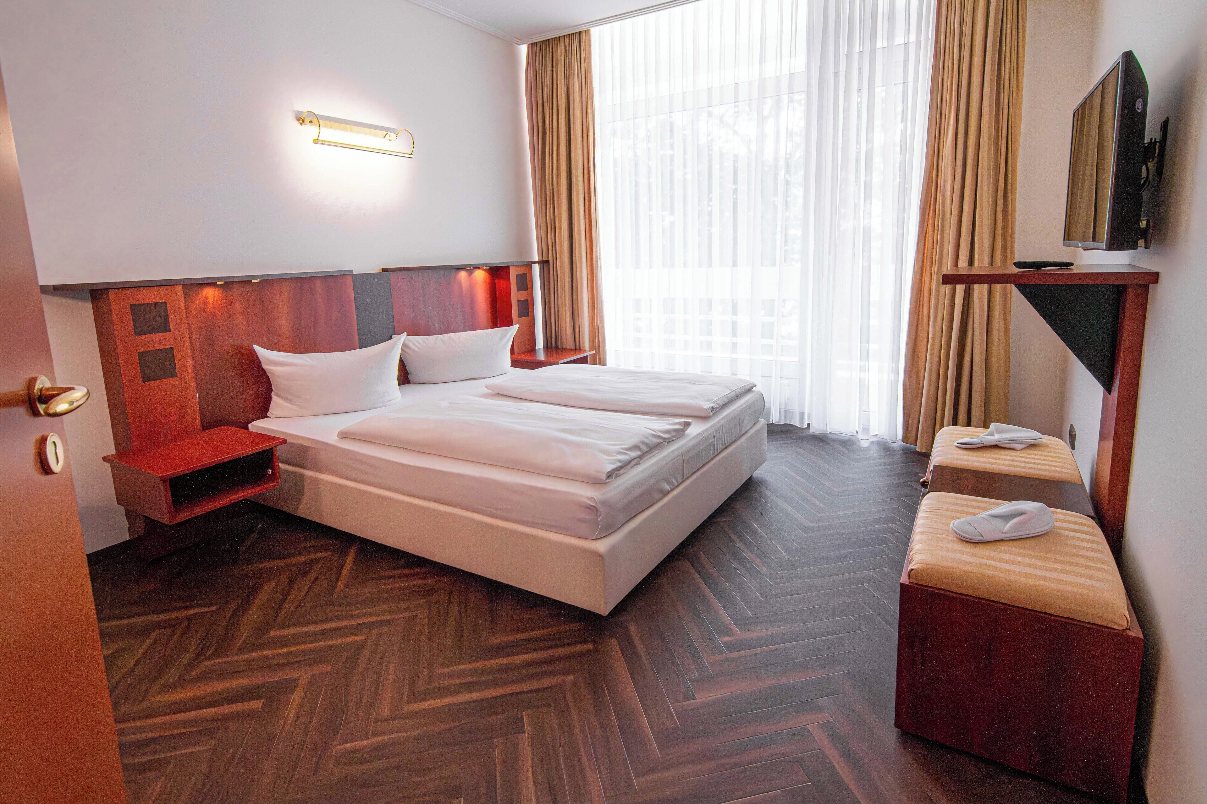 A hotel room with a large bed, a bedside table, a TV mounted on the wall, and a window with curtains.